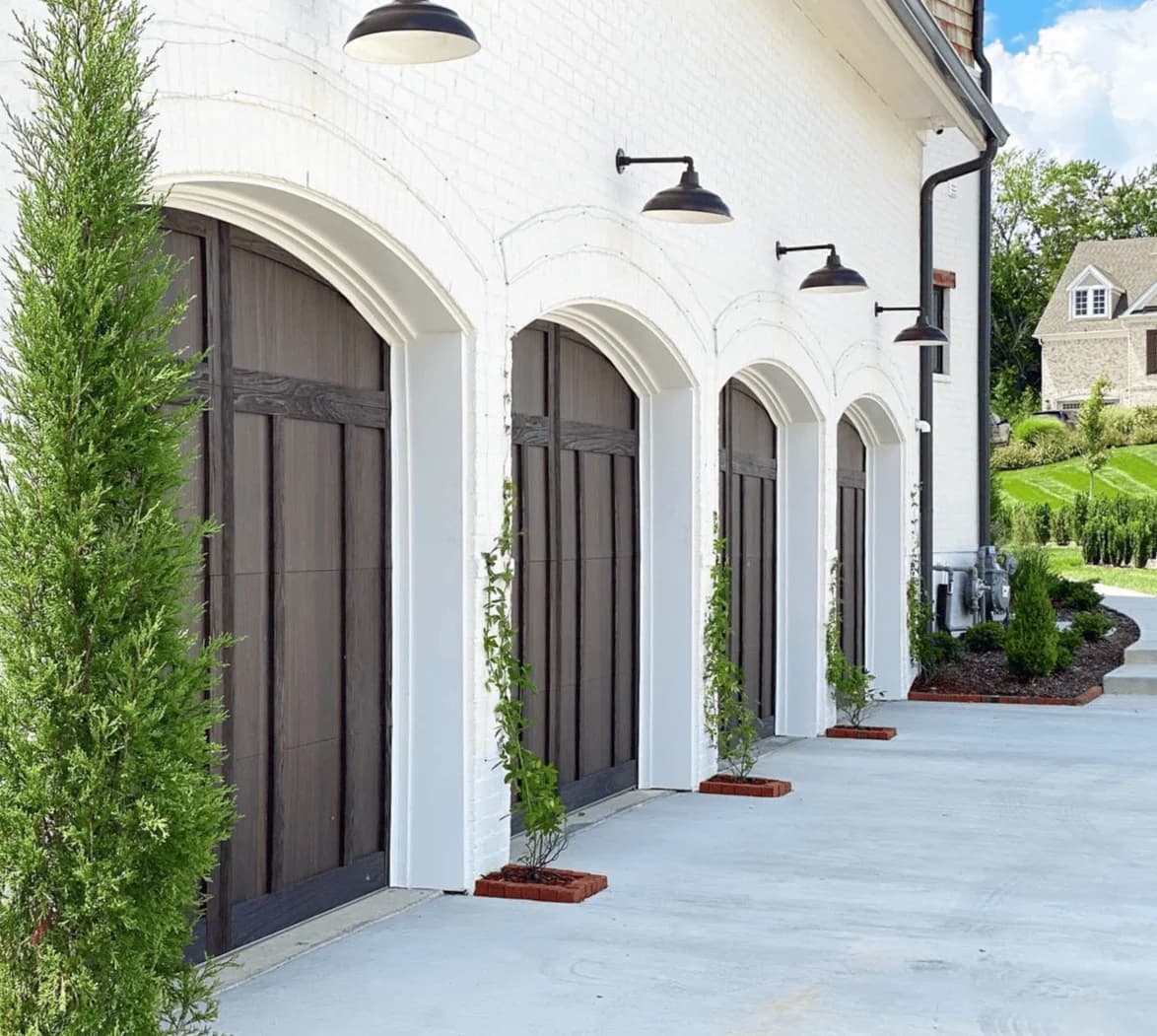 Row of arched dark wood-look garage doors on a white painted brick home with barn lights