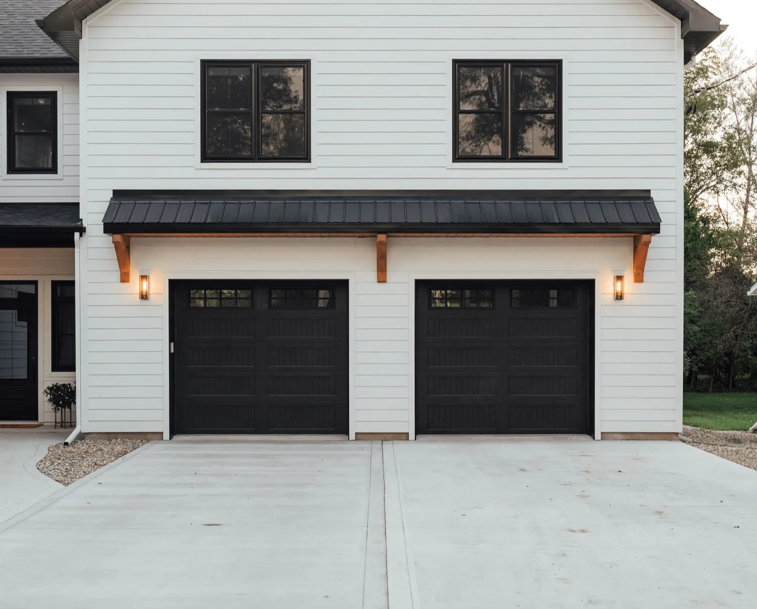 Black long-panel garage door under a metal awning on a white lap-sided home