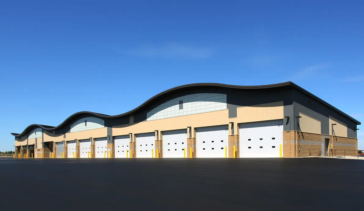 Row of white sectional steel garage doors along a large commercial building with a curved standing-seam roof and yellow bollards at each opening