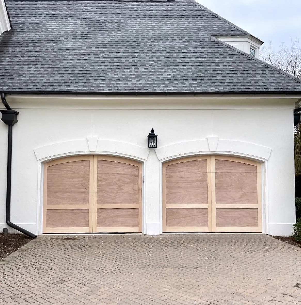 Two arch-top natural wood carriage-style garage doors on a white stucco home with a paver driveway
