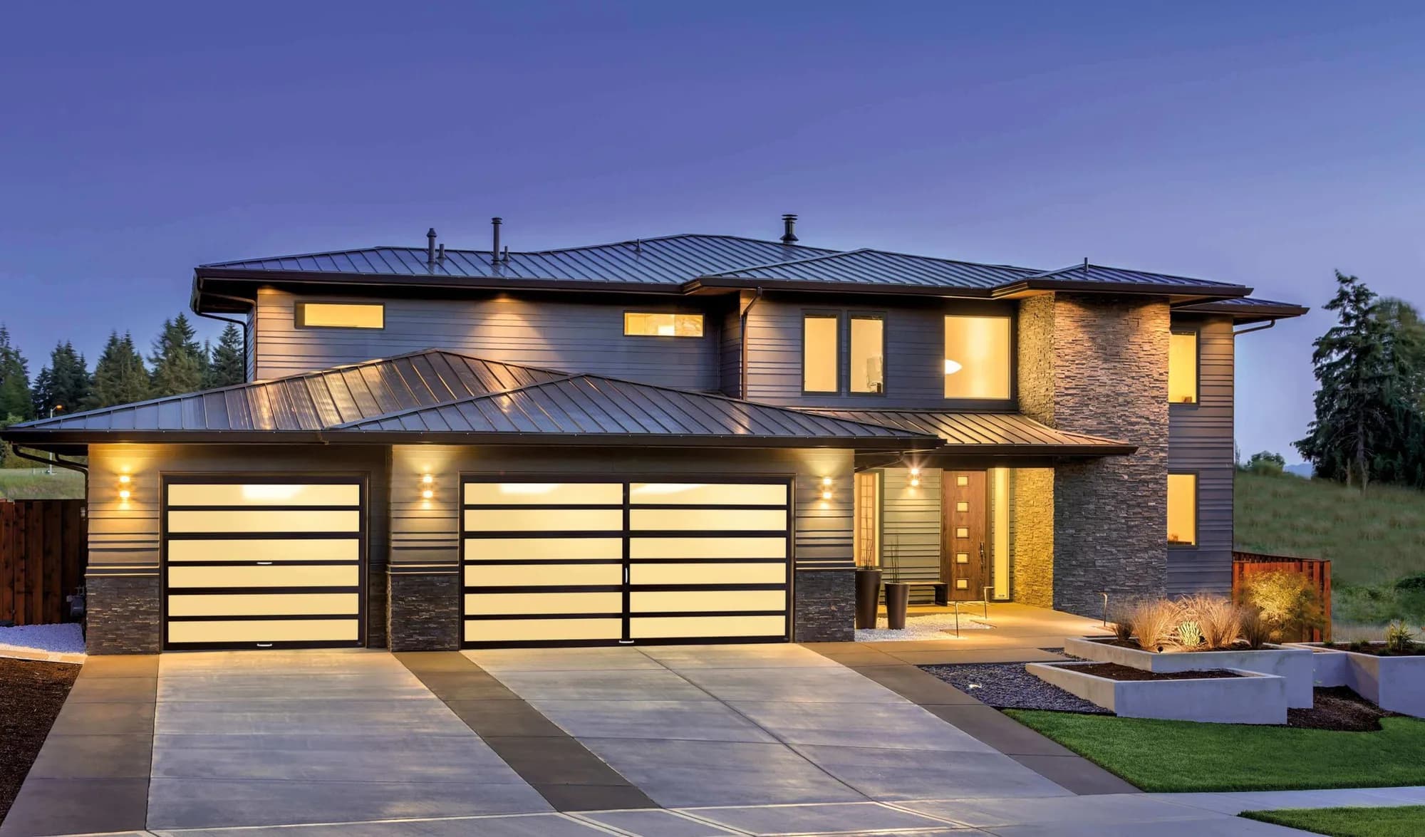 Three full-view garage doors with dark frames and frosted glass panels, lit at dusk on a modern two-story home