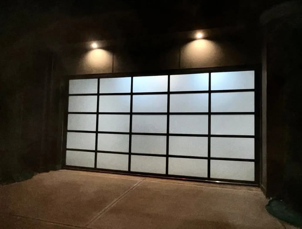 Modern full-view garage door with dark frame and frosted glass panels, lit by two overhead sconces at night
