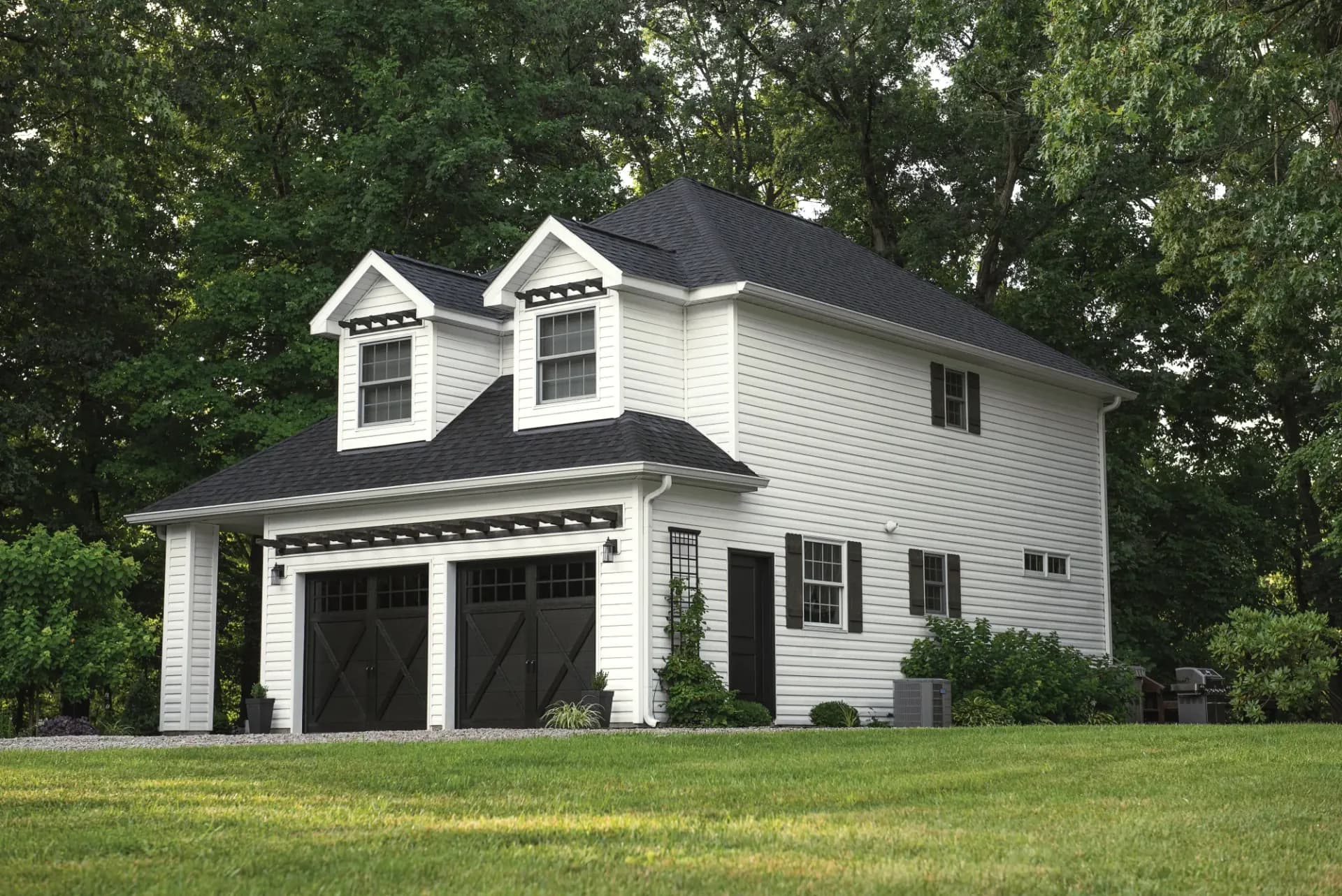 Two-story white carriage house with a black roof and two black carriage-style garage doors under a wood pergola, set against a wooded backdrop and green lawn