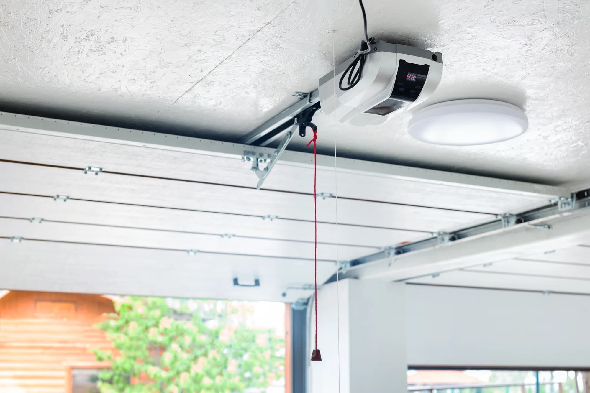 Ceiling-mounted garage door opener with its rail, trolley, and red emergency release cord above a white sectional door