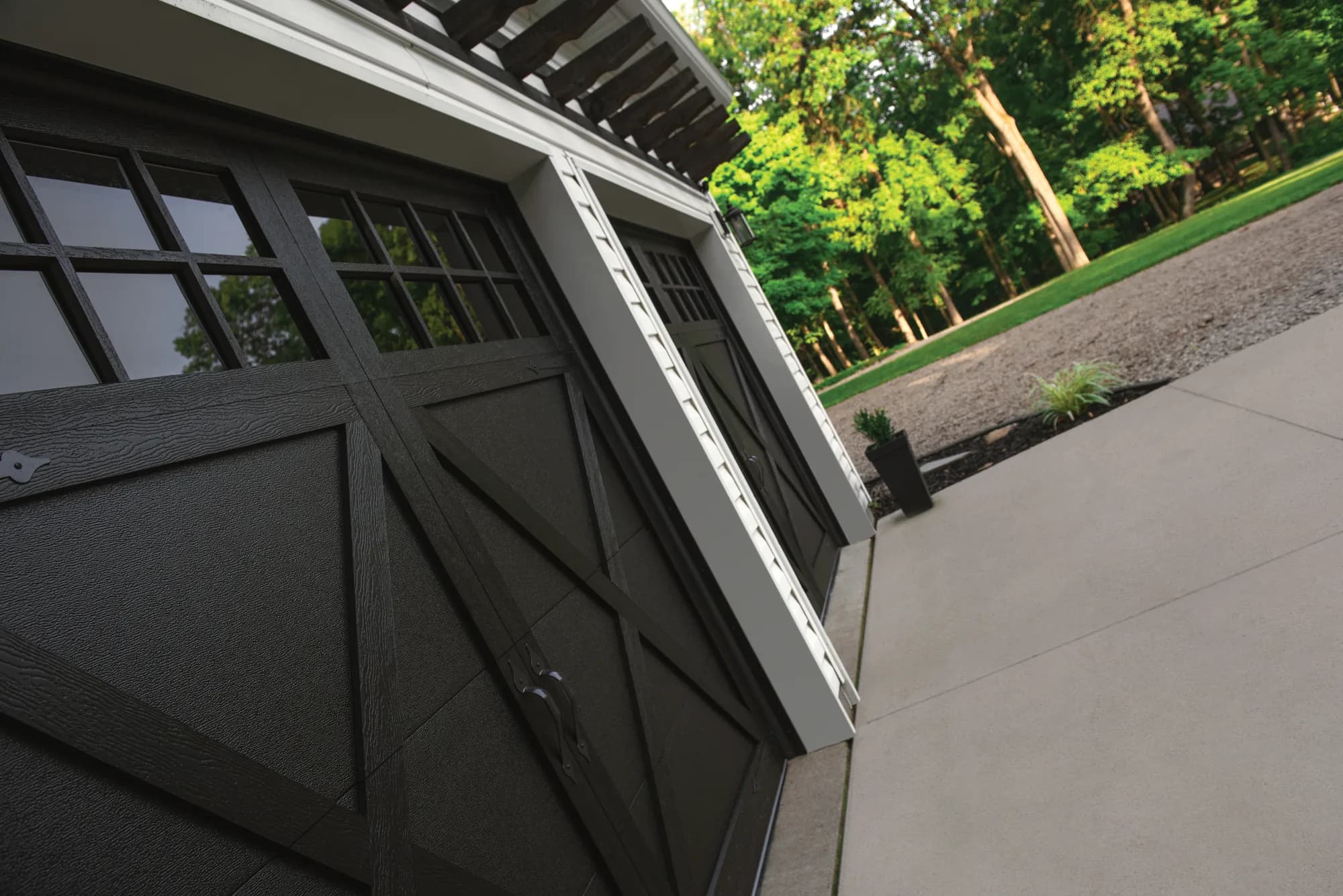 Close-up of a black carriage-house style garage door with X-brace detailing, divided-light windows, and decorative handles, beside a concrete driveway and treeline