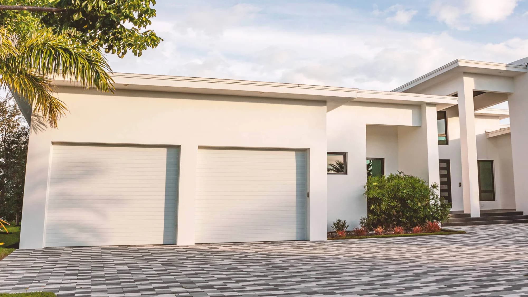 Modern white stucco home with two white flush-panel garage doors opening onto a paver driveway, framed by palms