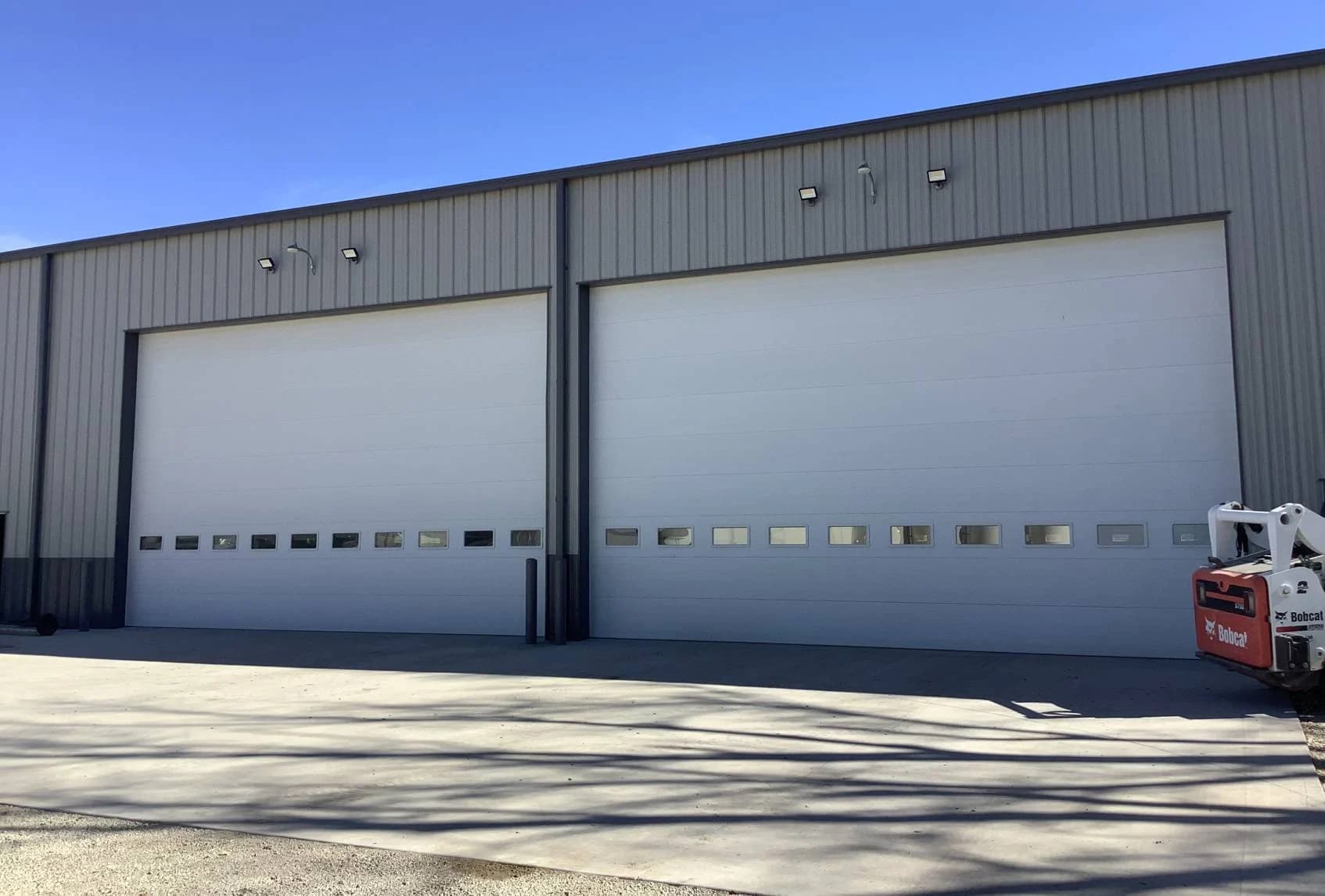 Two tall white insulated sectional steel doors with rows of vision panels on a grey metal shop building