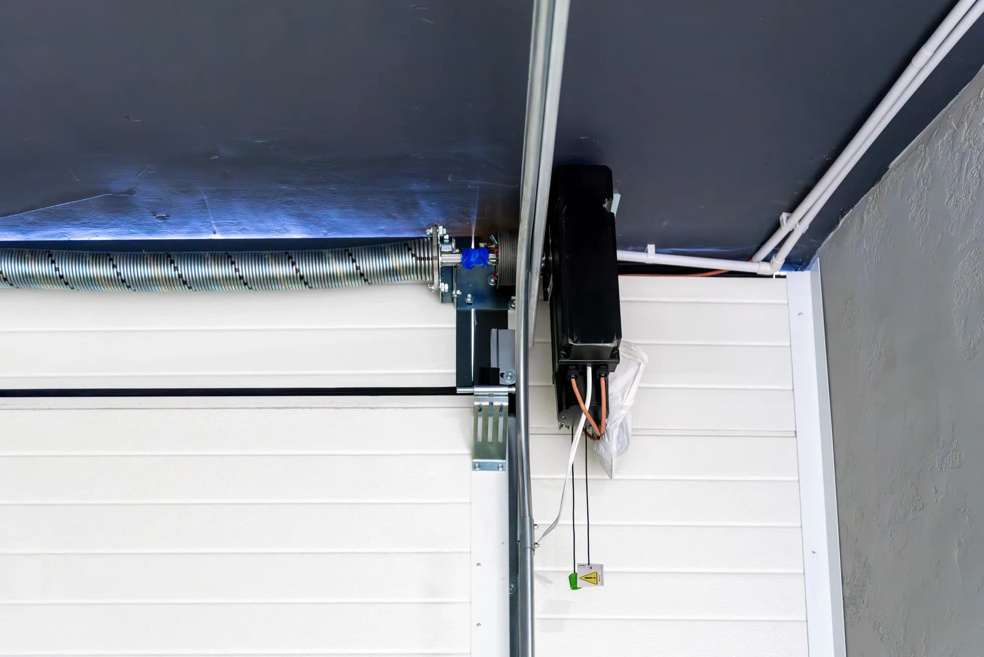 Vertical garage door track running down beside a white sectional door, with the torsion spring on its shaft and a side-mounted opener above